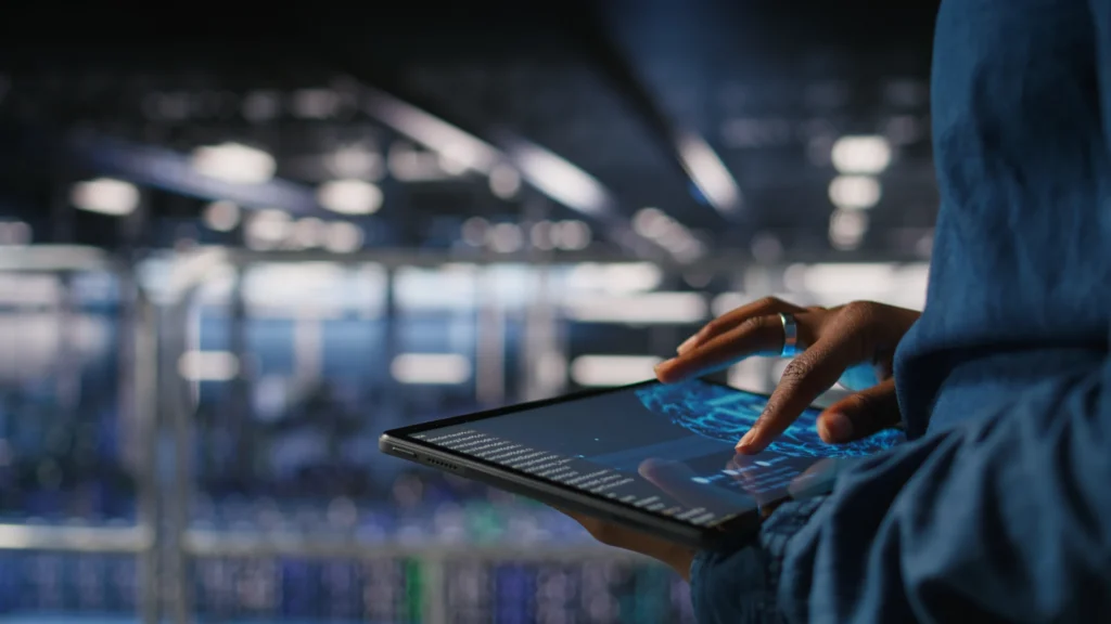 Close-up of a person’s hand tapping a tablet displaying a blue data chart, with a blurred data-center background in view.