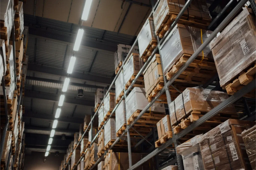 Warehouse aisle with tall metal racks and pallets of boxed goods wrapped in plastic, illuminated by overhead lights at night.